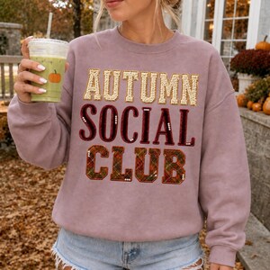 Peut inclure: Un sweat-shirt mauve à col rond avec le texte AUTUMN SOCIAL CLUB sur trois lignes. Le mot AUTUMN est en sequins dorés, SOCIAL est en velours rouge foncé, et CLUB est en tissu à carreaux vert et rouge avec de petites perles blanches.