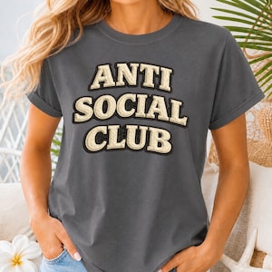 Peut inclure: Un t-shirt gris anthracite avec des manches retroussées. Le t-shirt affiche le texte "ANTI SOCIAL CLUB" en lettres majuscules crème avec un contour noir. Le t-shirt est de style décontracté et confortable.
