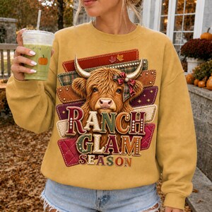Peut inclure: Un sweat-shirt à manches longues jaune moutarde avec un motif de vache Highland portant un nœud rouge sur la corne. Le design inclut des motifs rectangulaires superposés en tartan, cuir et textures matelassées avec le texte RANCH GLAM SEASON.