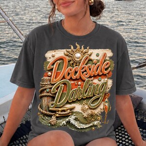 May include: Dark gray t-shirt with the words "Dockside Darling" in orange and green script. The graphic includes a sun, waves, and nautical elements like rope and a fishing lure. The shirt has short sleeves.
