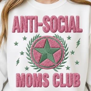 Op de afbeelding: Witte sweatshirt met roze geborduurde tekst "ANTI-SOCIAL MOMS CLUB". Een groene ster staat in het midden van een roze cirkel, omringd door groene laurierbladeren en sterren. Bliksemgraphics zijn ook aanwezig.