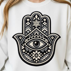 May include: White sweatshirt featuring a detailed embroidered Hamsa hand design. The black, gray, and white Hamsa hand includes an eye, floral elements, and geometric patterns. The sweatshirt is a comfortable, casual style.