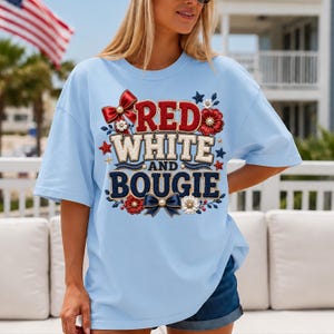 May include: Pale blue t-shirt featuring the text "RED WHITE AND BOUGIE" in red, white, and blue. The design includes bows, flowers, and stars. The shirt is a casual style.