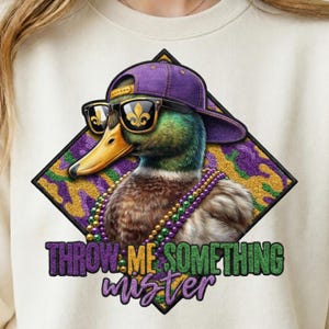 Peut inclure: Sweat-shirt crème avec un canard portant une casquette violette, des lunettes de soleil et un collier de perles. Le canard est placé sur un fond en forme de losange avec un motif camouflage violet, or et vert. Le texte dit "THROW ME SOMETHING mister."