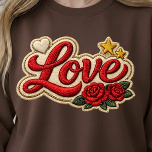Peut inclure: Sweat-shirt marron avec un motif brodé. Le motif comprend le mot "Love" en écriture rouge, un cœur blanc, deux étoiles jaunes et des roses rouges avec des feuilles vertes. Le motif est souligné en beige.