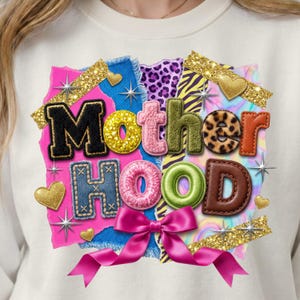 Peut inclure: Sweat-shirt blanc avec un motif "Motherhood" coloré. Le design présente un patchwork avec diverses textures, dont du denim, un imprimé léopard et des accents de paillettes dorées. Un nœud rose et des cœurs complètent le design.