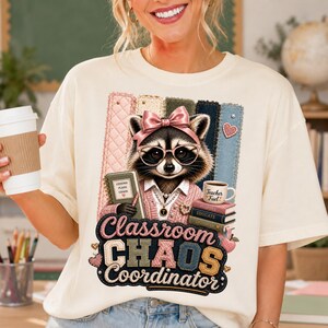 Peut inclure: Un t-shirt crème avec un motif de raton laveur portant des lunettes et un nœud rose, vêtu d'un gilet. Le raton laveur tient un carnet près d'une pile de livres marqués 'Educate' et 'Empower' et d'une tasse avec l'inscription 'Teacher Fuel'.