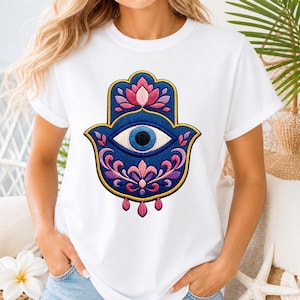 Peut inclure: T-shirt blanc orné d'une broderie détaillée représentant une main de Hamsa. Le motif comprend un œil bleu, une fleur de lotus rose et des motifs floraux ornés dans des tons de rose, de violet et d'or. La main de Hamsa est un symbole de protection.