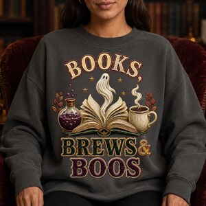 Peut inclure: Un sweat-shirt à col rond gris anthracite avec une impression graphique portant le texte "BOOKS, BREWS & BOOS". Le motif représente un livre ouvert avec un fantôme s'élevant des pages, une fiole de potion violette, une tasse fumante et des étoiles dorées.