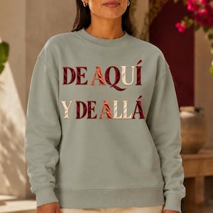 Peut inclure: Un sweat-shirt à col rond vert sauge arborant le texte brodé DE AQUÍ Y DE ALLÁ dans un dégradé de tons rouge foncé, orange et crème. Le lettrage est disposé sur deux lignes avec une fine ligne décorative incurvée reliant les mots.