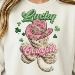 Peut inclure: Sweat-shirt crème avec un chapeau de cow-boy rose orné de trèfles, le mot "Lucky" en vert et "Cowgirl" en rose. Le chapeau est posé sur un trèfle en corde, parfait pour la Saint-Patrick.