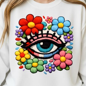 Könnte beinhalten: Weißes Sweatshirt mit einem farbenfrohen Stickdesign. Das Design zeigt ein großes Auge mit blauer Iris, umgeben von leuchtenden Blumen in Rot, Blau, Gelb, Rosa, Grün und Lila. Das Auge hat schwarze Wimpern und ein rosa-schwarzes Augenlid.