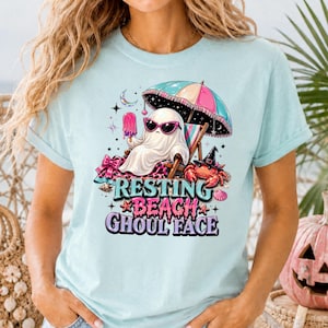 Op de afbeelding: Lichtblauw T-shirt met een afbeelding van een spook met een zonnebril, ontspannen op een strandstoel onder een roze en turquoise parasol, met een ijsje. De tekst op het shirt luidt "Resting Beach Ghoul Face."