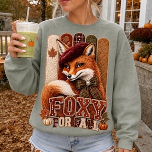 Peut inclure: Un sweat-shirt col rond vert sauge avec un motif de renard portant un béret bordeaux et un nœud papillon à carreaux. Le design comprend des panneaux texturés verticaux, des feuilles d'automne, des citrouilles et le texte FOXY FOR FALL.