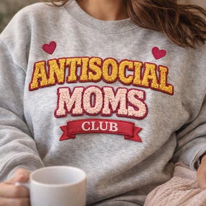 May include: A light gray sweatshirt with the text "ANTISOCIAL MOMS CLUB" in a retro style. The text is in yellow and pink with red accents, and two red heart patches are above the text. A white mug is held in the person's hand.