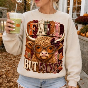 Peut inclure: Un sweat-shirt beige à col rond orné d'un motif de vache Highland portant des lunettes à imprimé léopard et un nœud bordeaux. Le texte indique FALL BUT FANCY, entouré de panneaux verticaux texturés orange, verts et marron avec des feuilles d'automne.