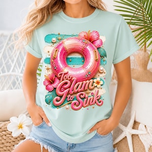 Könnte beinhalten: Hellblaues T-Shirt mit einem farbenfrohen Aufdruck. Es zeigt einen rosa-orangenen Schwimmring mit dem Text "Too Glam to Sink". Das Design umfasst Hibiskusblüten, Perlen und goldfarbene Akzente.