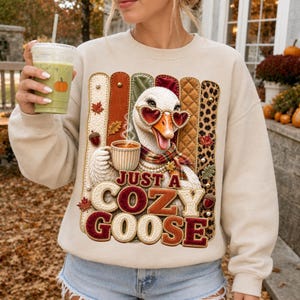 Peut inclure: Un sweat-shirt beige à col rond orné d'une oie portant des lunettes de soleil en forme de cœur et une écharpe, tenant une tasse de café. Le motif inclut des bandes texturées, des feuilles d'automne, des glands et le texte "JUST A COZY GOOSE".