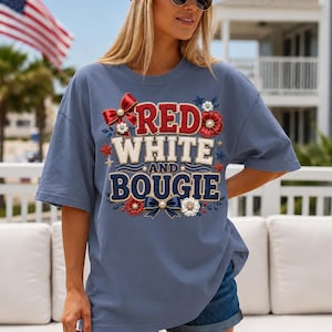 Peut inclure: Un t-shirt bleu ardoise à manches courtes avec un motif graphique portant le texte RED WHITE AND BOUGIE. Le design comprend des lettres style broderie rouge, blanc et bleu, des nœuds décoratifs, des fleurs et de petites étoiles patriotiques.