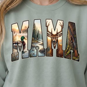 May include: A light green sweatshirt with the word "MAMA" in large, block letters. Each letter features a different outdoor scene: a duck on a lake, a forest, a deer, and a rifle. The design has a hunting or outdoors theme.