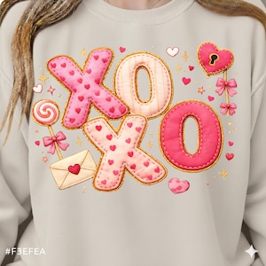 Peut inclure: Un sweat-shirt beige clair avec un motif de Saint-Valentin. Le motif comprend les lettres "XO" en rose et crème, décorées de cœurs. Des éléments supplémentaires incluent un cadenas en forme de cœur, une sucette et une enveloppe.