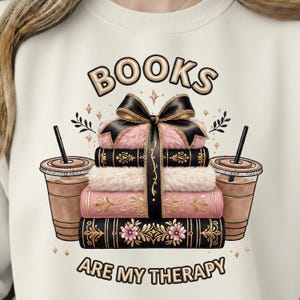 Puede incluir: Sudadera color crema con una pila de libros, un lazo negro, piel rosa y detalles dorados. El texto "BOOKS ARE MY THERAPY" está debajo, con dos cafés helados a cada lado.