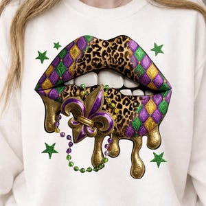 May include: White sweatshirt featuring a graphic of stylized lips with a leopard print center, and purple, green, and gold harlequin pattern. A gold fleur-de-lis and dripping gold accents are included, along with green star accents.