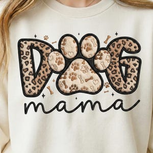 May include: Cream-colored sweatshirt with a graphic design featuring the word "DOG" in a leopard print pattern, a paw print design, and the word "mama" in a cursive font. The paw print design includes bones and hearts.
