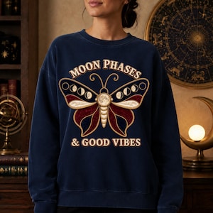 Peut inclure: Un sweat-shirt bleu marine à col rond orné d'un motif de papillon dont les ailes représentent les phases de la lune. Le texte "MOON PHASES & GOOD VIBES" est imprimé en couleur crème au-dessus et au-dessous de l'illustration du papillon.