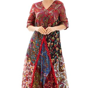May include: A colorful, patterned maxi dress with a V-neck and elbow-length sleeves. The dress features a patchwork design with paisley and floral prints in red, blue, and green. A red sun hat completes the look.