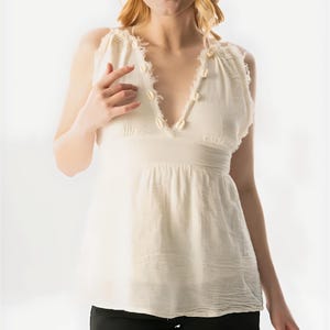May include: Cream-colored sleeveless top with a V-neckline, adorned with small seashells. The top has a relaxed fit and a textured appearance. Paired with black denim shorts. The model is blonde.