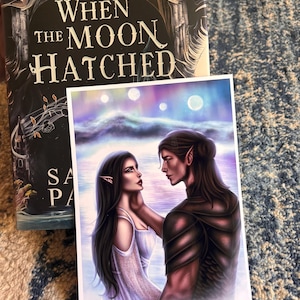 May include: A book titled "When the Moon Hatched" with a fantasy art print. The print depicts two figures with long dark hair and pointed ears, set against a celestial background. The book cover has a dark, detailed design.