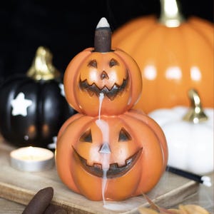 May include: A stacked orange jack-o'-lantern incense burner featuring two carved pumpkin faces. A lit incense cone sits on top of the upper pumpkin, releasing a thick white smoke that flows downward through the mouths of both carved pumpkins.
