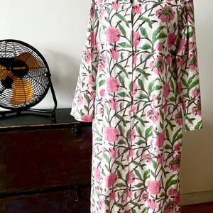 May include: A long, white dress with a floral pattern of pink flowers and green leaves. The dress has long sleeves and a button-up front. It is displayed on a black mannequin head.