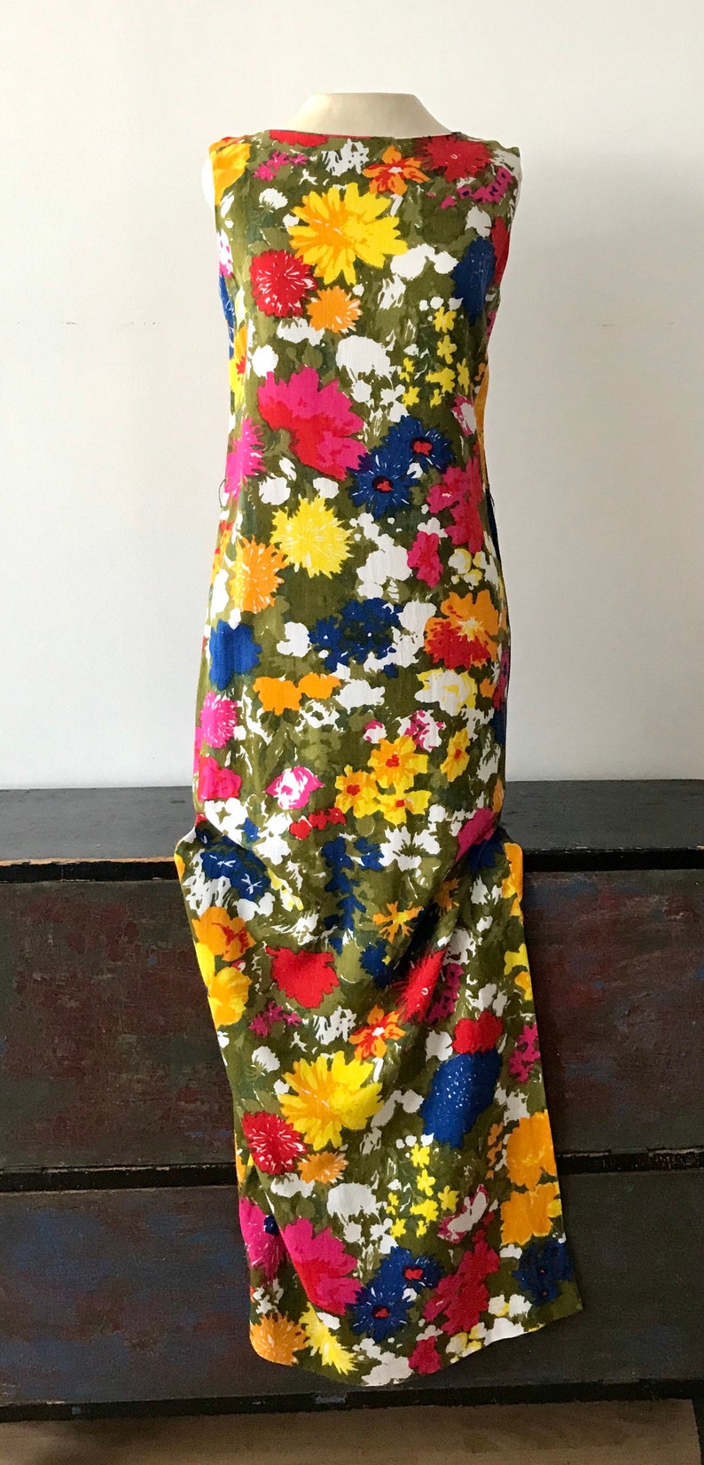 May include: A sleeveless maxi dress with a vibrant floral print. The dress features a variety of colors, including yellow, red, pink, blue, and green. The flowers are large and bold, and the dress has a simple, elegant silhouette.