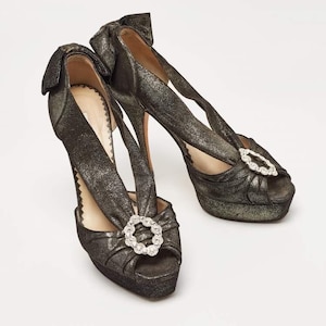 May include: A pair of metallic bronze high-heeled shoes with open toes. Each shoe features a bow at the heel and a decorative rhinestone buckle. The platform heels are approximately 1 inch high, and the stiletto heels are approximately 4 inches high.