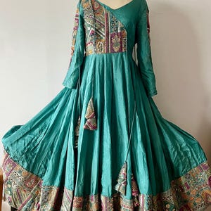 May include: A teal-colored, long-sleeved dress with intricate embroidery and embellishments. The dress features a V-neckline, a flowing skirt, and decorative tassels. The neckline is adorned with a matching beaded necklace.