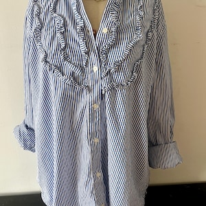 May include: Blue and white striped button-down shirt with ruffled trim. The long-sleeved shirt has a mandarin collar and is buttoned down the front. The sleeves are rolled up. A necklace is around the collar.