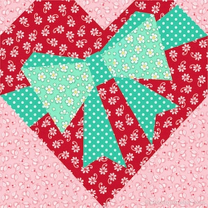 Op de afbeelding: Een rood hartvormig quiltblok met een groene strik in het midden. De strik is gemaakt van verschillende stoffen met patronen, waaronder een groene stof met witte stippen en een groene stof met witte bloemen.