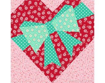 May include: A red heart-shaped quilt block with a green bow in the center. The bow is made of different patterned fabrics, including a green fabric with white polka dots and a green fabric with white flowers.