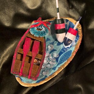 Puede incluir: Una escena náutica decorativa en miniatura con un bote rojo y marrón con remos, una pequeña corona azul y dos boyas. La escena está sobre una base azul y dorada con detalles transparentes en forma de gema.