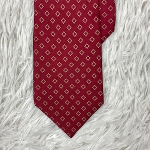 Peut inclure: Une cravate rouge foncé ornée d'un motif répétitif de petits losanges blancs. La cravate présente une extrémité pointue et une texture tissée lisse. Le design géométrique se compose de minuscules points blancs formant le contour de chaque losange.