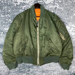 Peut inclure: Un blouson aviateur vert olive doté d'une fermeture éclair frontale intégrale, d'un col, de poignets et d'un ourlet côtelés. La veste comprend deux poches avant à boutons-pression et une poche utilitaire sur la manche gauche. La doublure est orange.