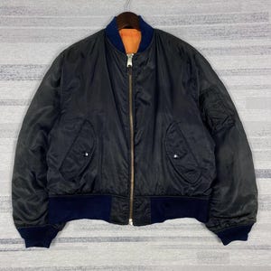 May include: A black bomber jacket featuring a central metal zip, a dark blue ribbed collar, cuffs, and hem. The jacket includes two front pockets with press-stud closures, a utility pocket on the left sleeve, and a bright orange interior lining.