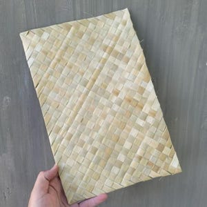 Puede incluir: Un mantel individual rectangular tejido con paja natural de color claro. La paja está tejida en un patrón diagonal, creando una superficie texturizada. El mantel individual está sostenido por una mano sobre un fondo gris.