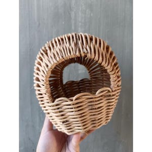 May include: A small, round woven basket with a handle. The basket is made of light brown wicker and has a scalloped edge. The handle is a circular opening at the top. The basket is held by a hand.
