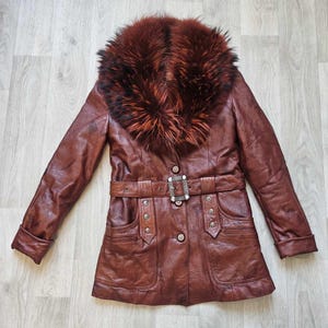 May include: A brown leather jacket with a fur collar. The jacket has a belted waist with a decorative buckle, two front pockets, and button closures. The fur collar is a mix of brown and reddish-brown tones.