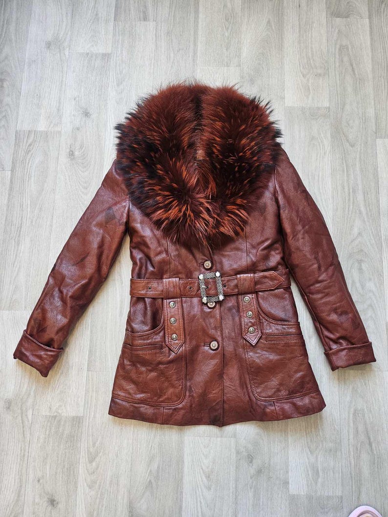 May include: A brown leather jacket with a fur collar. The jacket has a belted waist with a decorative buckle, two front pockets, and button closures. The fur collar is a mix of brown and reddish-brown tones.