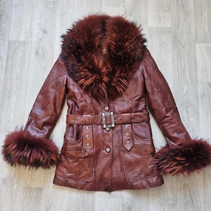 May include: A brown leather coat with a fur collar and cuffs. The coat features a belted waist with a decorative buckle and button closures. The fur trim is a mix of brown and reddish-orange.