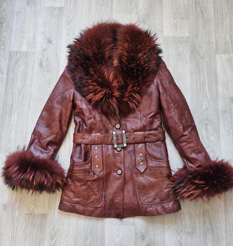 May include: A brown leather coat with a fur collar and cuffs. The coat features a belted waist with a decorative buckle and button closures. The fur trim is a mix of brown and reddish-orange.